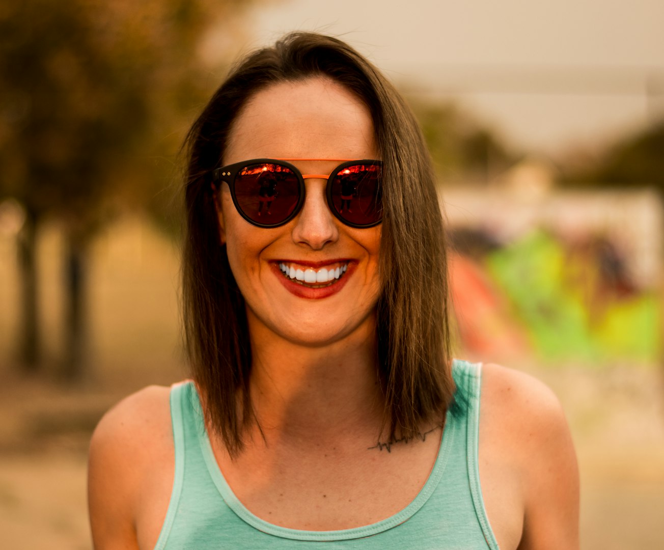 A smiling young woman with shoulder-length dark hair is standing in a bright, modern room with light-colored furniture and minimal decor. She is wearing a plain white t-shirt and blue jeans.